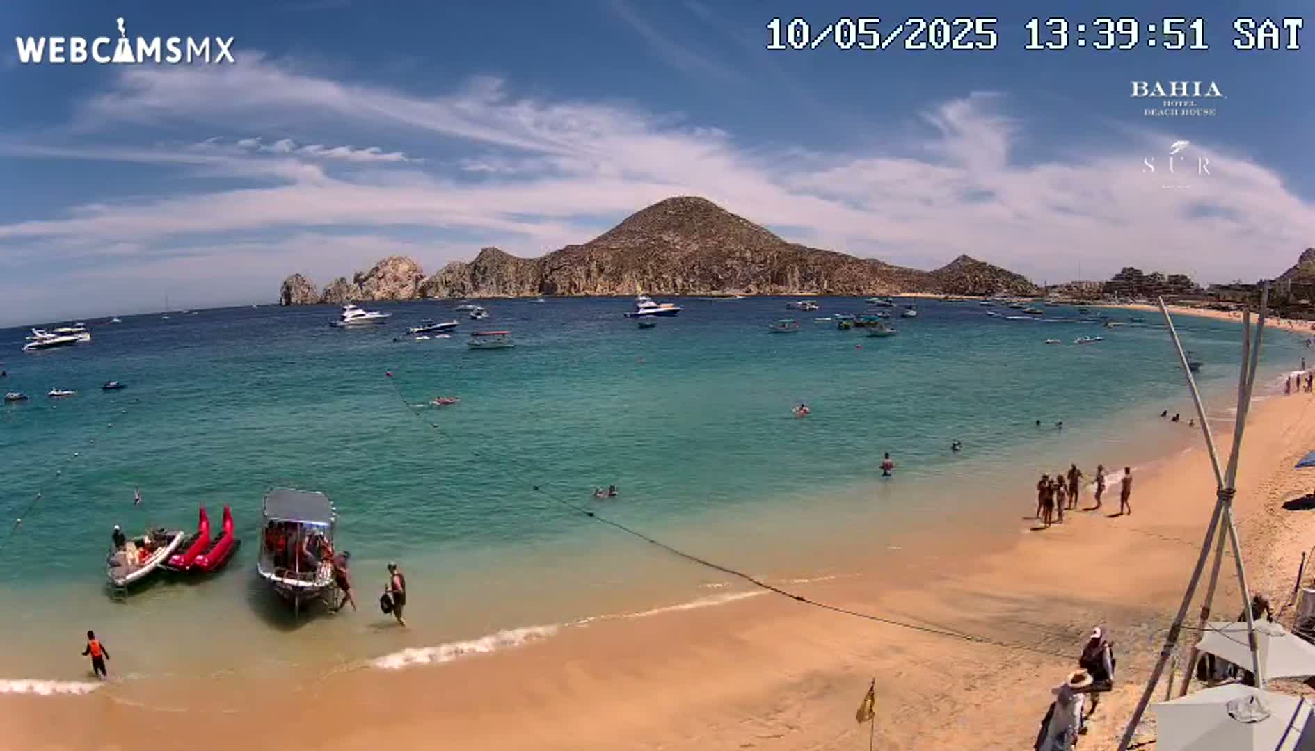 A sandy beach with turquoise water, several boats, and people swimming and sunbathing under a mostly sunny sky.