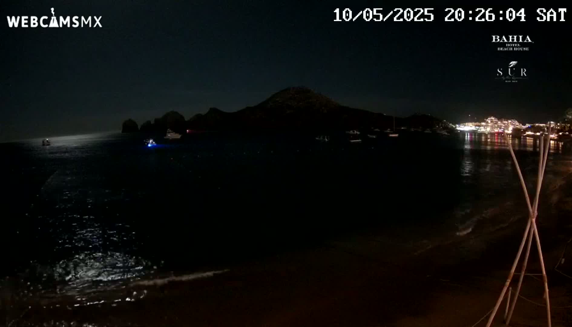 A dark nighttime beach scene shows a calm body of water reflecting a dark silhouette of land with a few lights visible in the distance.