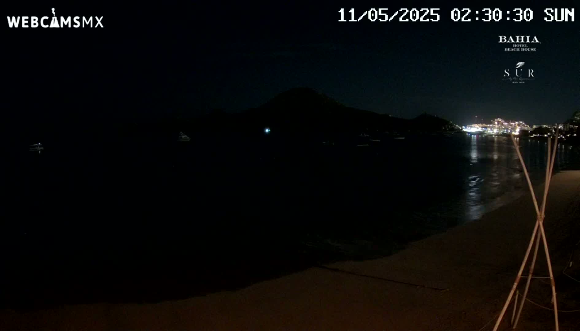A dark, nighttime beach scene shows a calm body of water reflecting distant lights, with a hill silhouetted against the night sky.