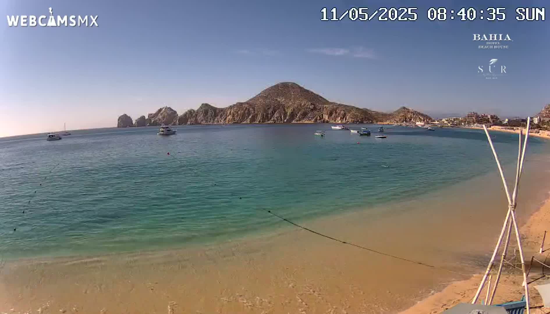 A sandy beach with calm, clear turquoise water stretches out to a bay with several boats, backed by rocky hills under a sunny sky.