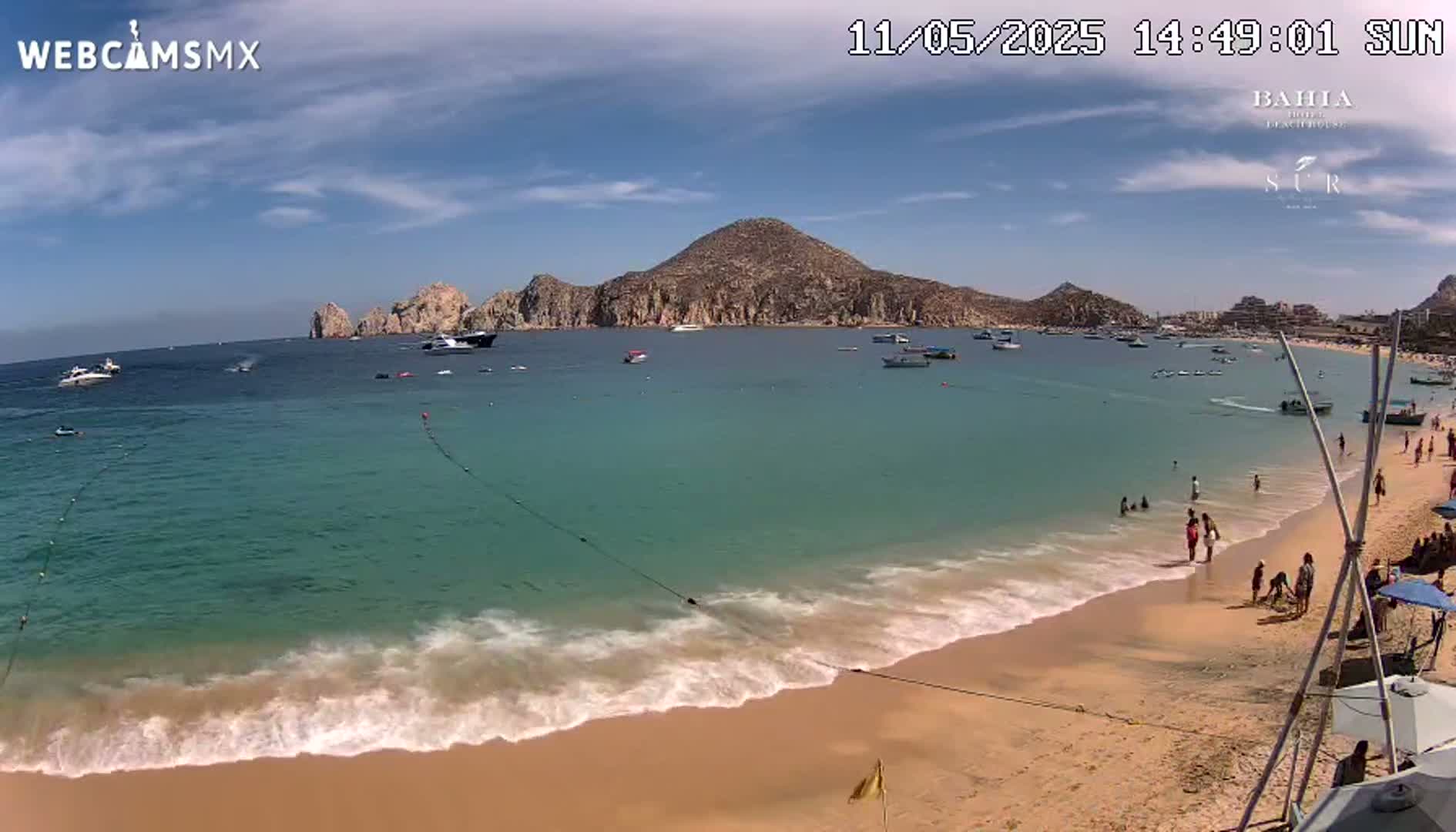A sandy beach with calm, turquoise water laps gently at the shore under a sunny sky, with several boats visible in the bay and people enjoying the beach.