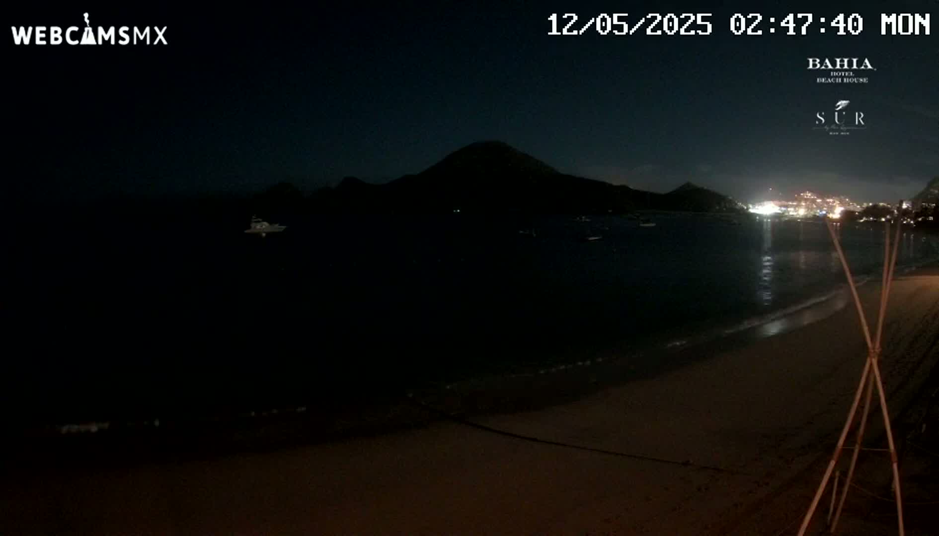 A dark nighttime beach scene shows a calm body of water, a distant mountain silhouette, and a few faint lights on the opposite shore.
