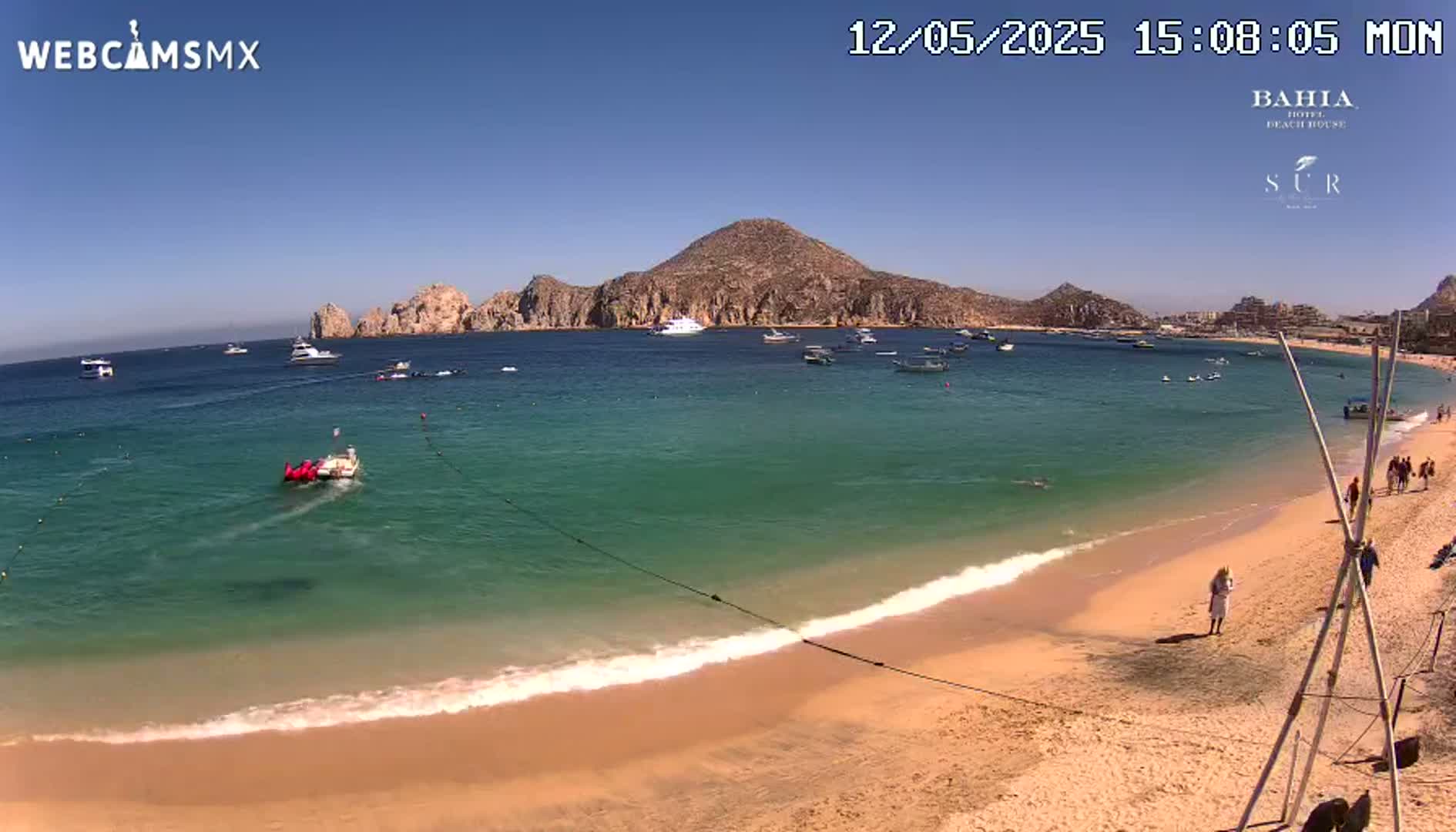 A sandy beach with turquoise water laps gently at the shore under a clear blue sky, with boats and people visible in the distance and a few people on the beach.
