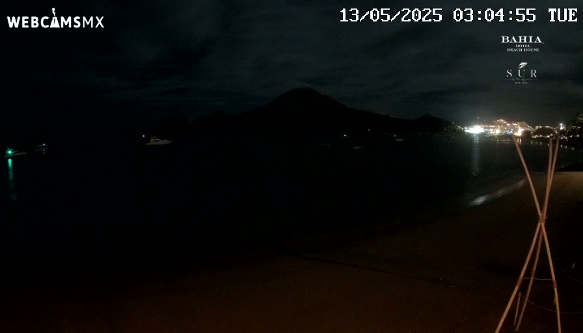 A dark night scene shows a mostly black body of water, a dark mountain in the distance, and a dimly lit beach with a tall, thin, wooden structure in the foreground.