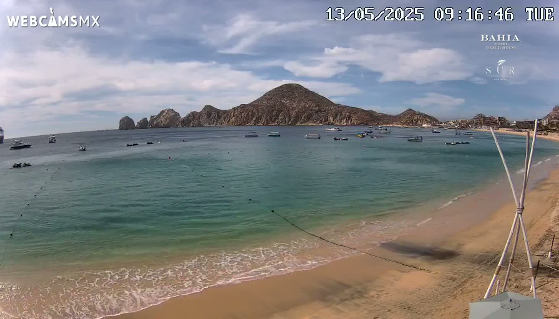 A sandy beach with calm, clear turquoise water, several boats, and rocky mountains in the background under a partly cloudy sky.