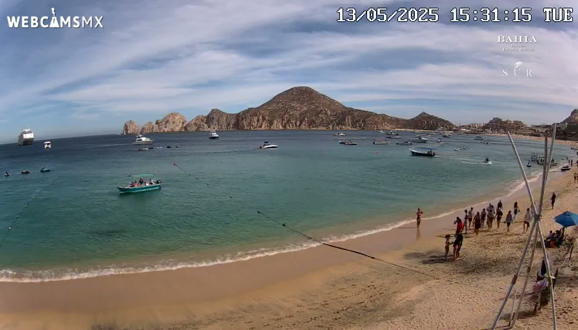 A sandy beach with calm, turquoise water, several boats, a large cruise ship in the distance, and a group of people walking along the shore under a mostly sunny sky.
