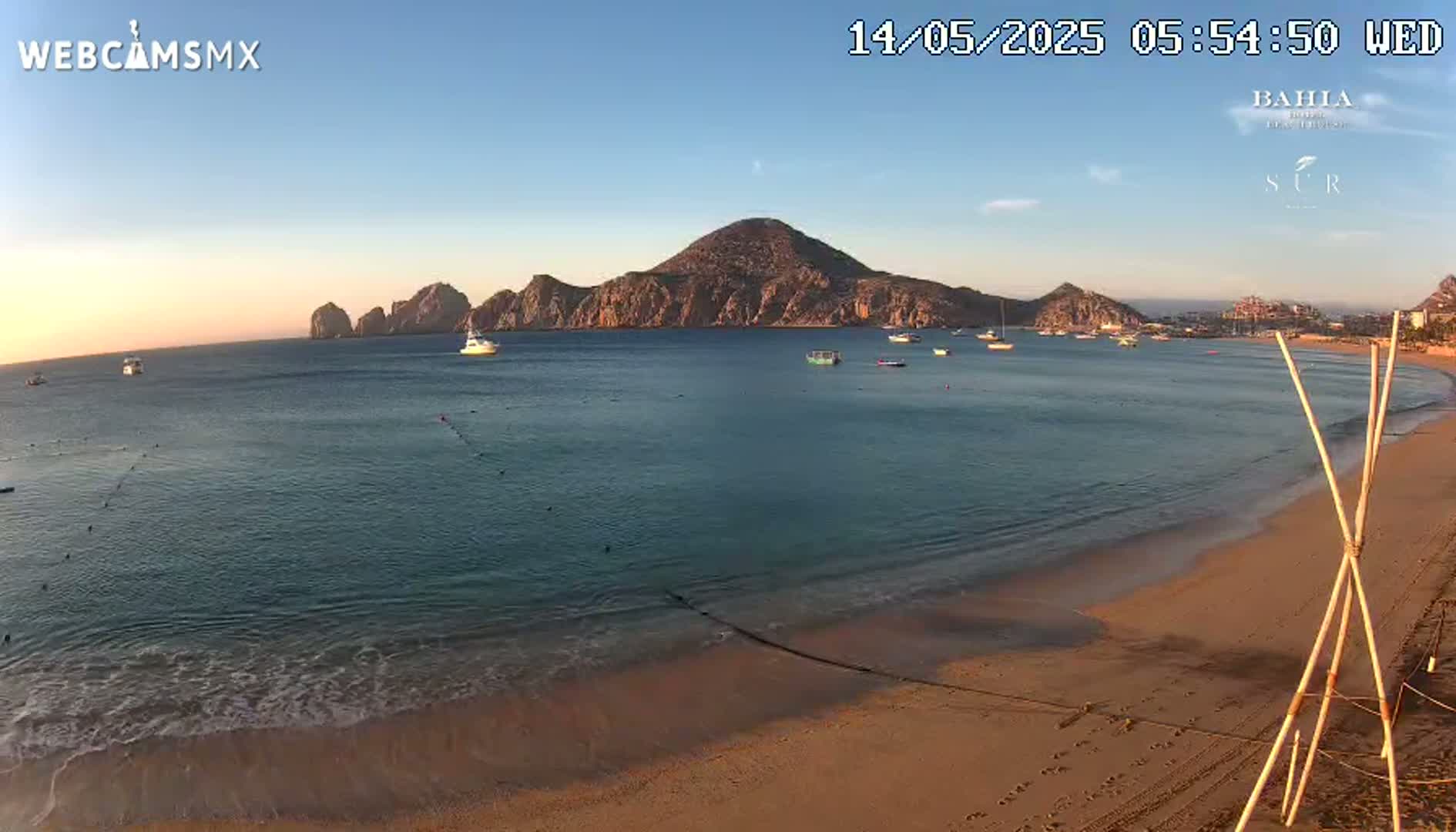 A sandy beach with calm ocean waters and several boats, under a clear sunset sky, features a rocky island in the distance.