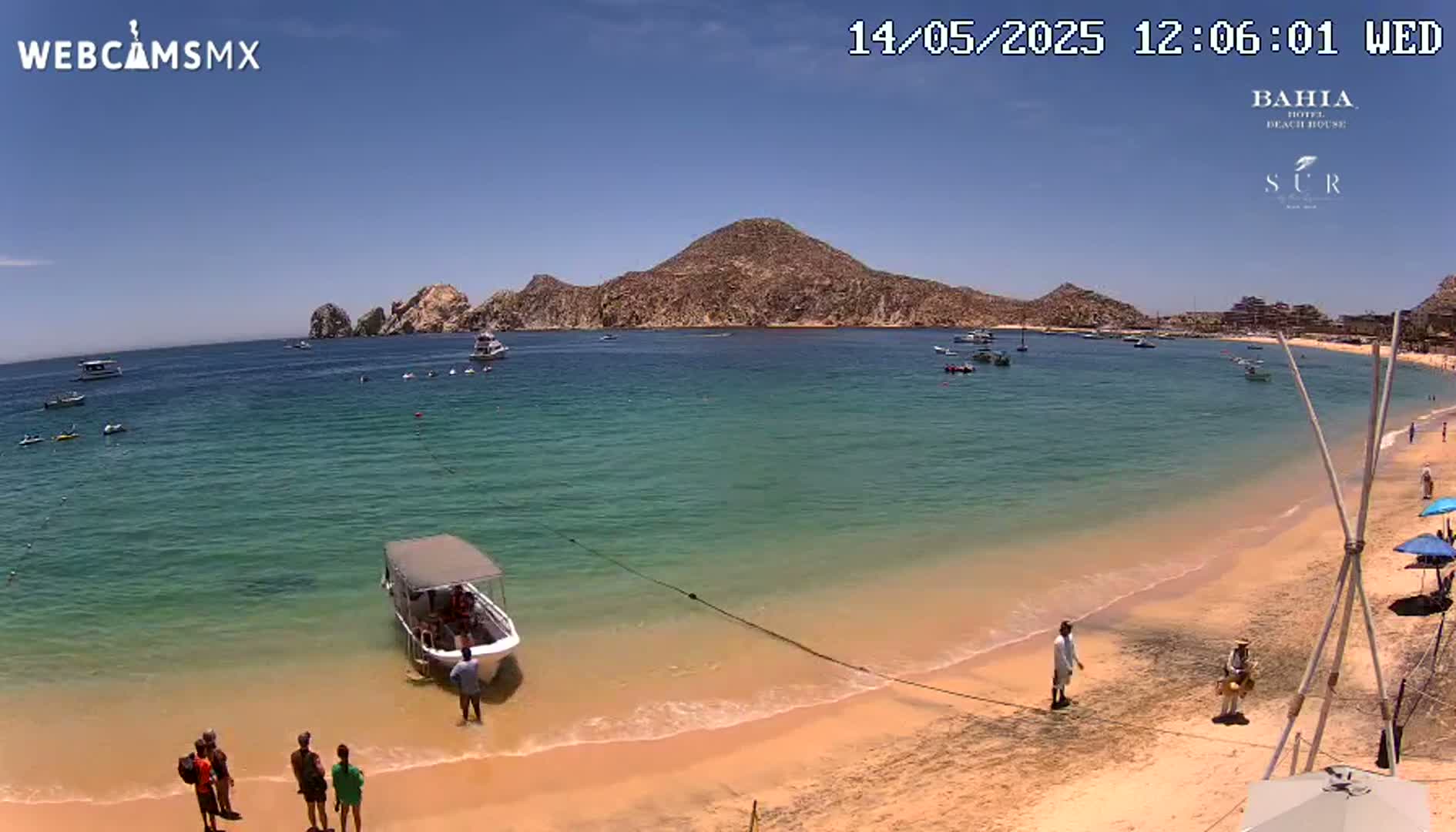 A sunny beach with clear, turquoise water, several boats, and people on the shore near a rocky hill in the background.