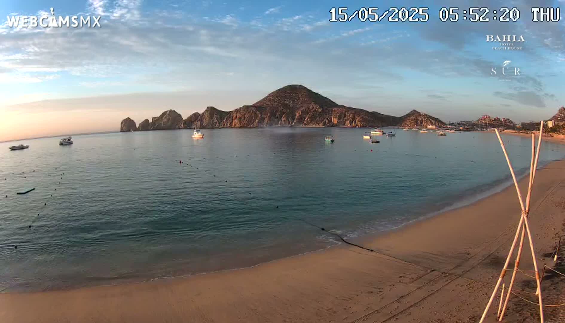 A sandy beach at sunrise, calm water with several boats, and a mountainous peninsula in the background.