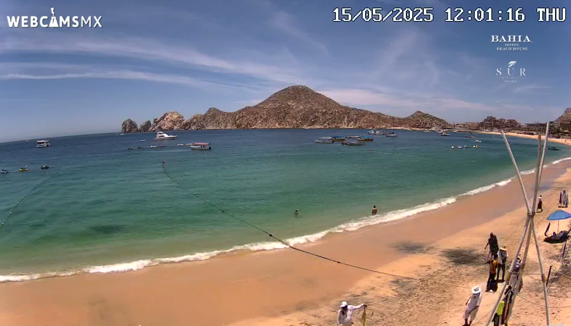 A sandy beach with calm, clear turquoise water laps gently against the shore, under a sunny sky with a few clouds, and a large rocky hill in the background.