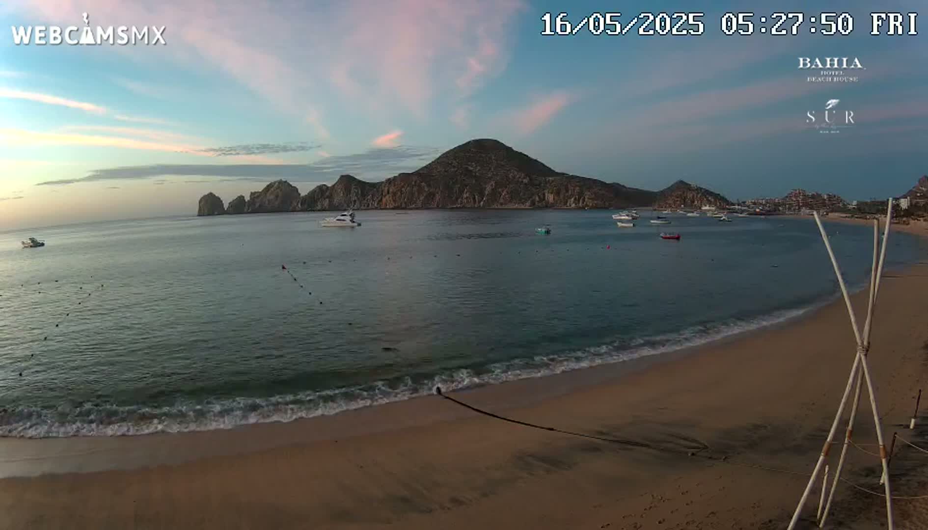 A sandy beach with gentle waves laps the shore under a clear, sunrise sky, overlooking a calm bay with several boats and a mountainous island in the distance.