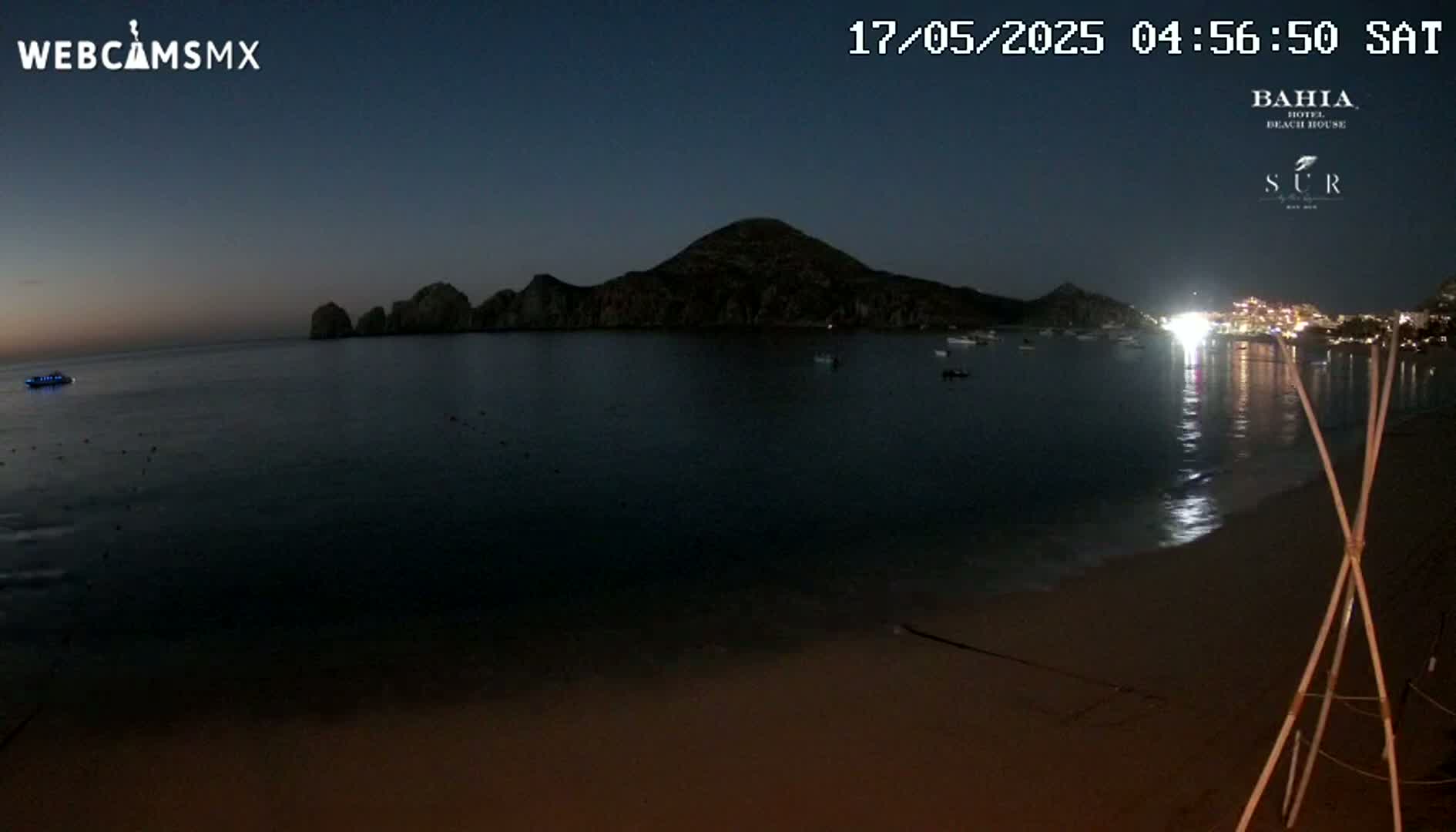 A dark, calm bay at twilight is seen from a beach, with a large hill or mountain in the background and lights visible on the opposite shore.
