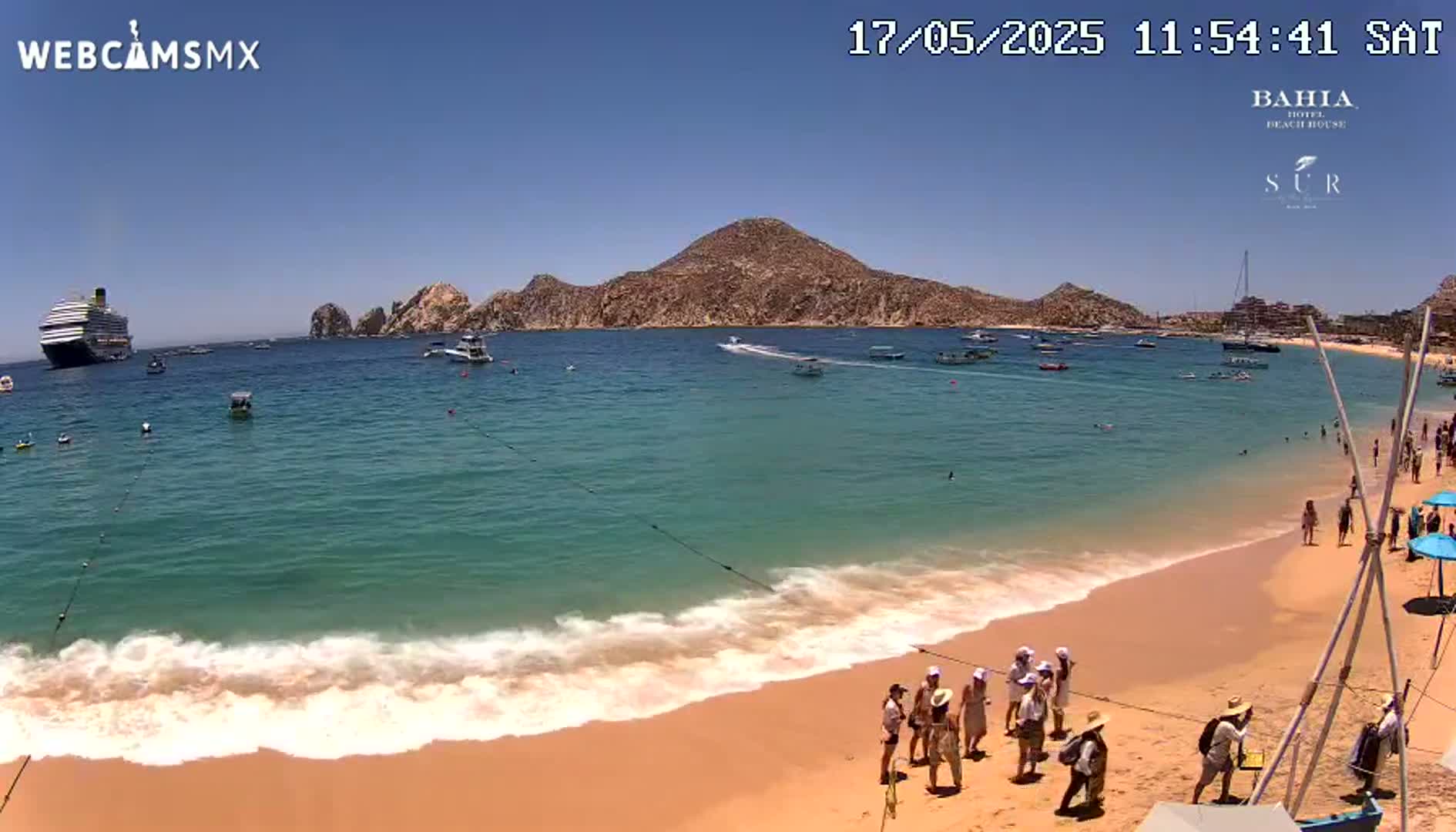 A sandy beach with gentle waves laps the shore under a clear blue sky, with a large cruise ship and several smaller boats visible in the turquoise water beyond, and a mountainous landscape in the background.