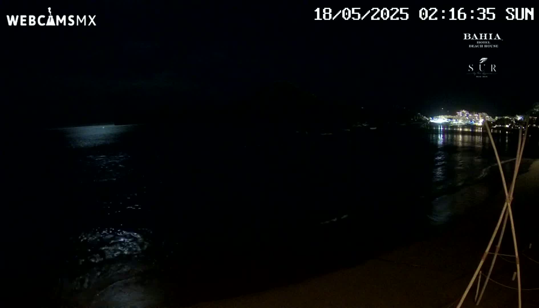 A dark nighttime beach scene shows a calm ocean reflecting the lights of a town across the water.