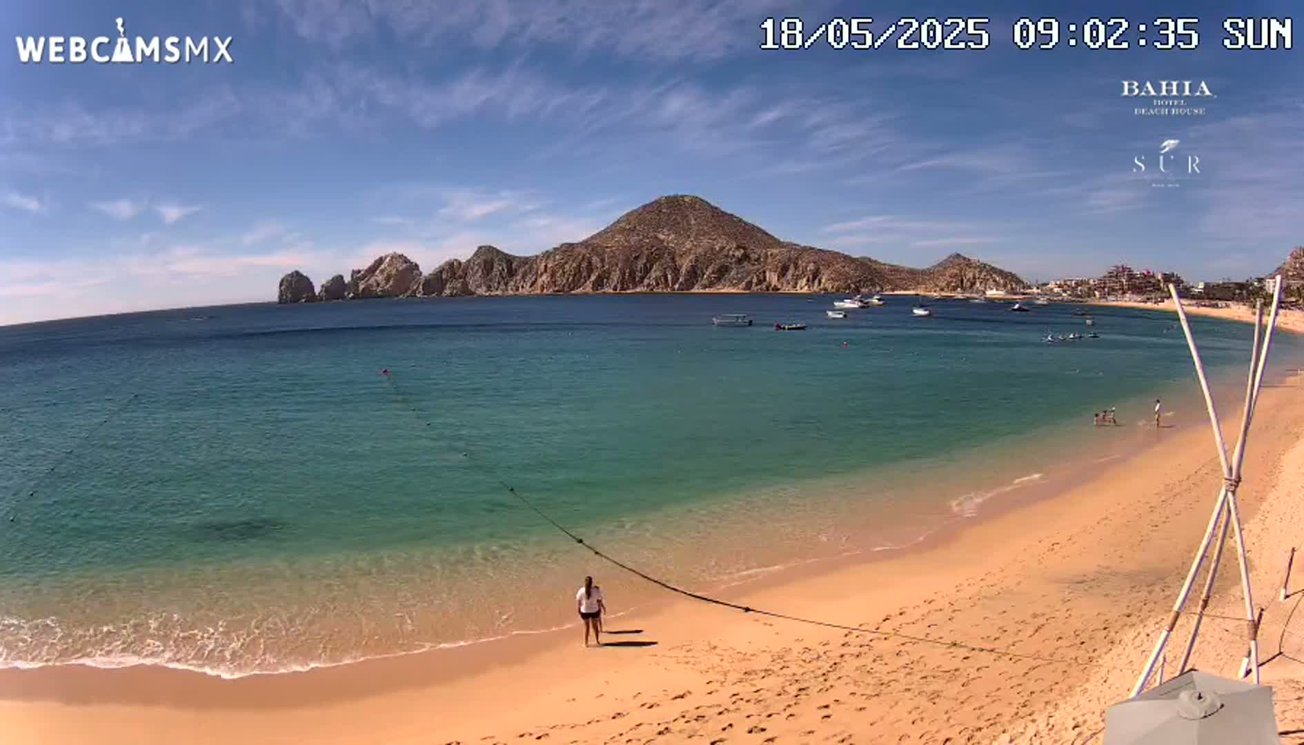 A sandy beach with clear, turquoise water laps gently at the shore, under a mostly sunny sky, with a large, rocky peninsula in the background.