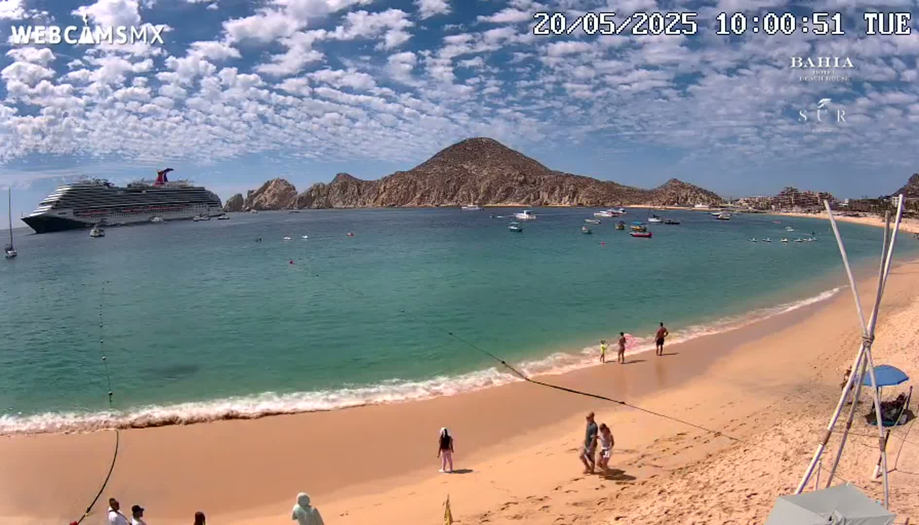 A large cruise ship is anchored in a calm bay near a sandy beach with several people on it under a mostly sunny sky with scattered clouds.