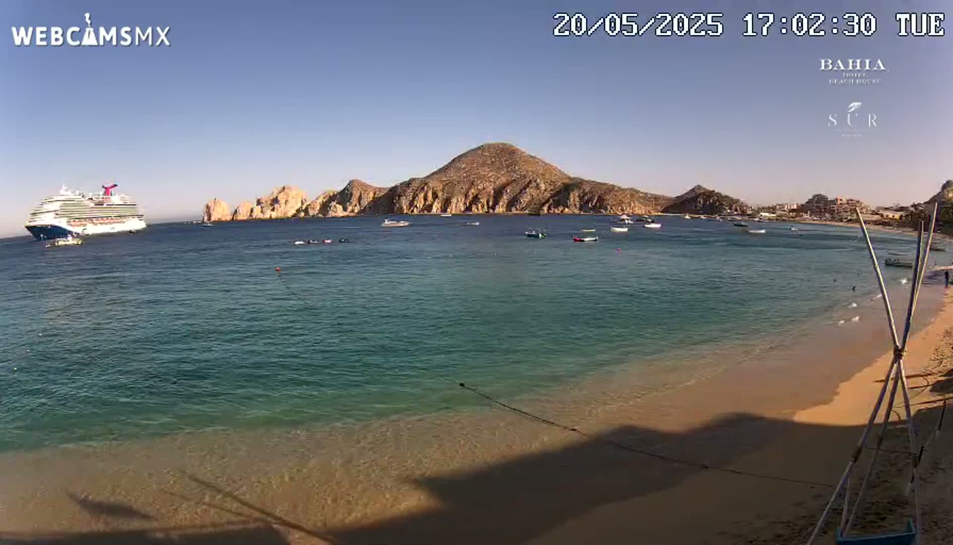 A cruise ship is anchored offshore from a sandy beach and a rocky island, under sunny skies.