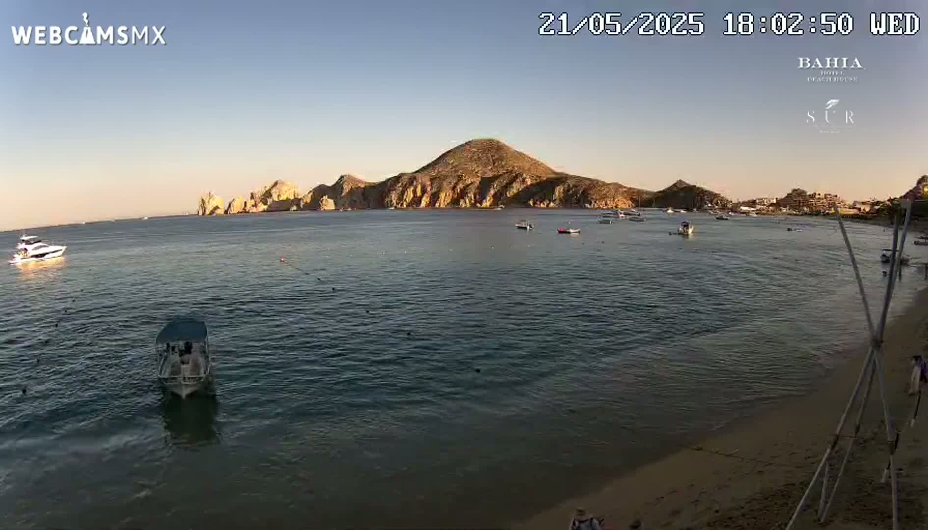 A calm, sunlit ocean bay shows several boats, a sandy beach, and a large rocky island in the background.