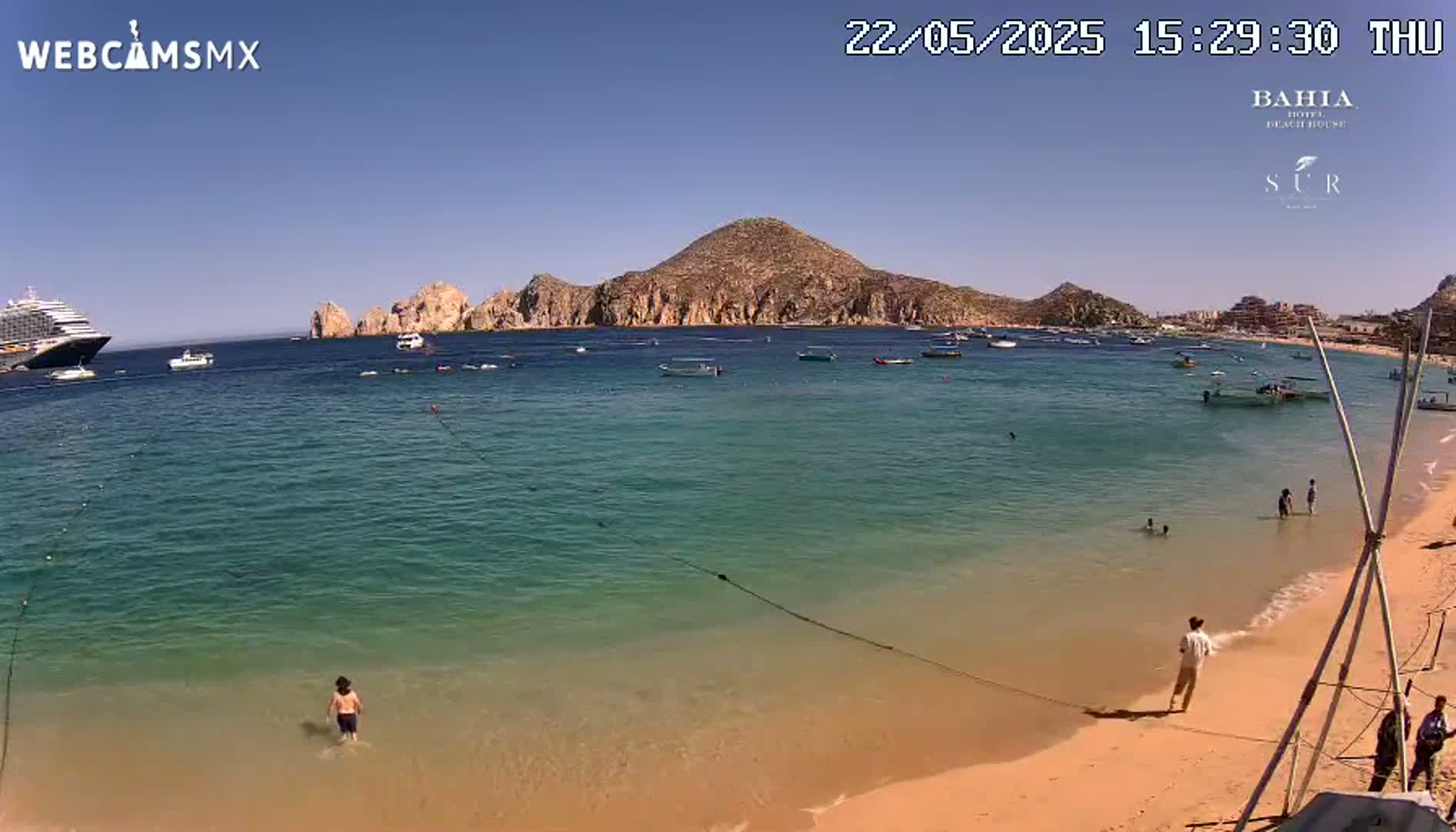 A sandy beach with calm, clear turquoise water, a few people swimming and walking on the shore, several small boats on the water, a large cruise ship in the distance, and a mountainous landscape under a clear blue sky.