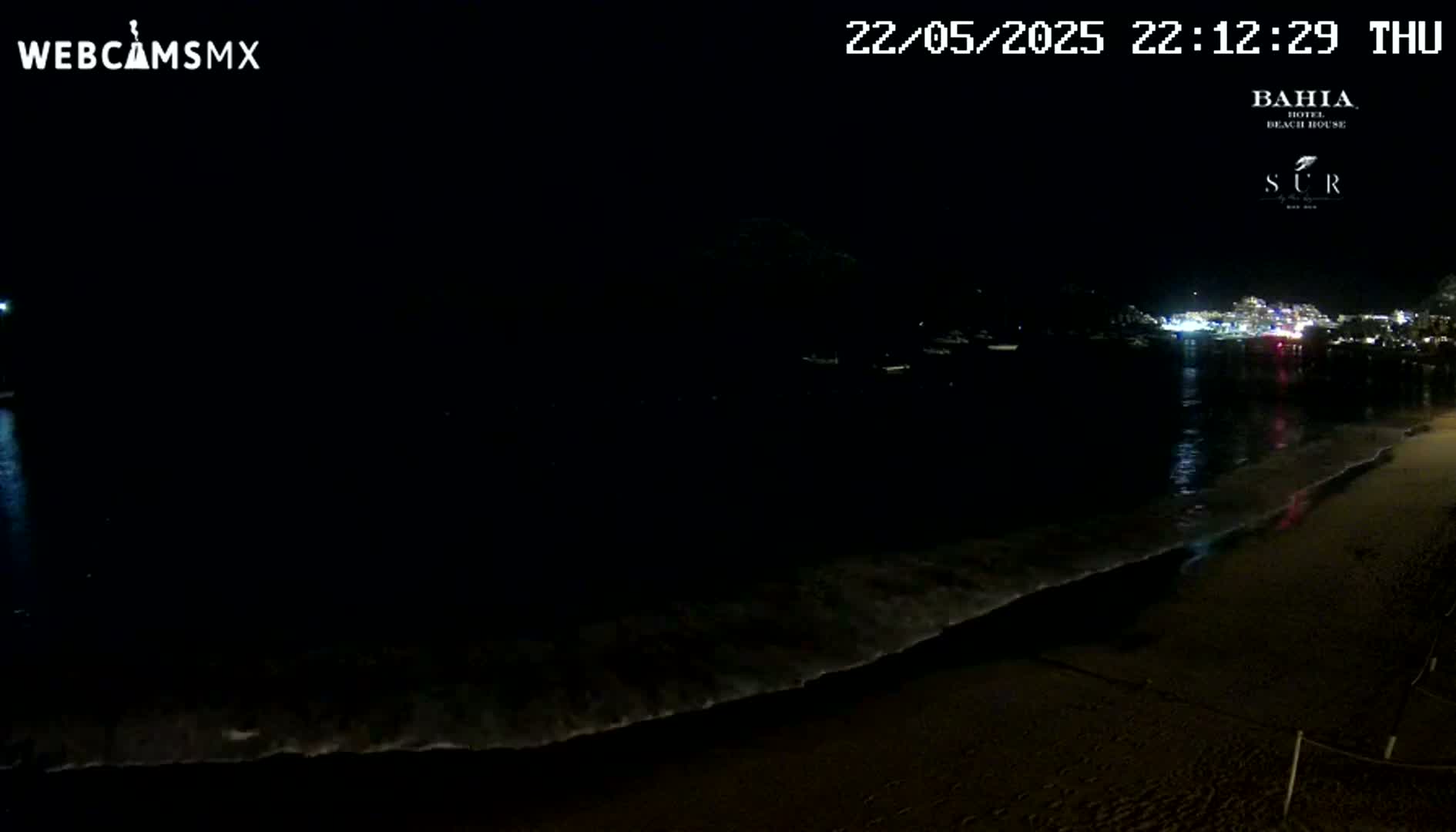 A dark, nighttime beach scene shows gentle waves lapping on the shore, with faintly lit buildings visible across the water.