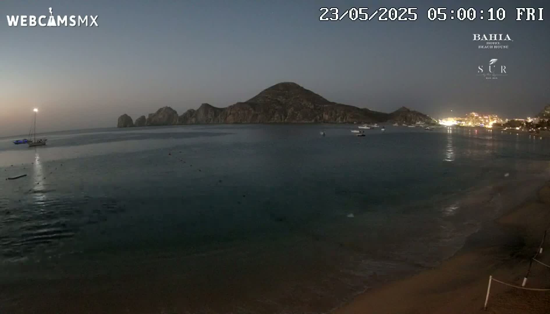 A tranquil nighttime beach scene shows calm, dark water lapping a sandy shore under a dusky sky, with a distant, mountainous island and a few boats.