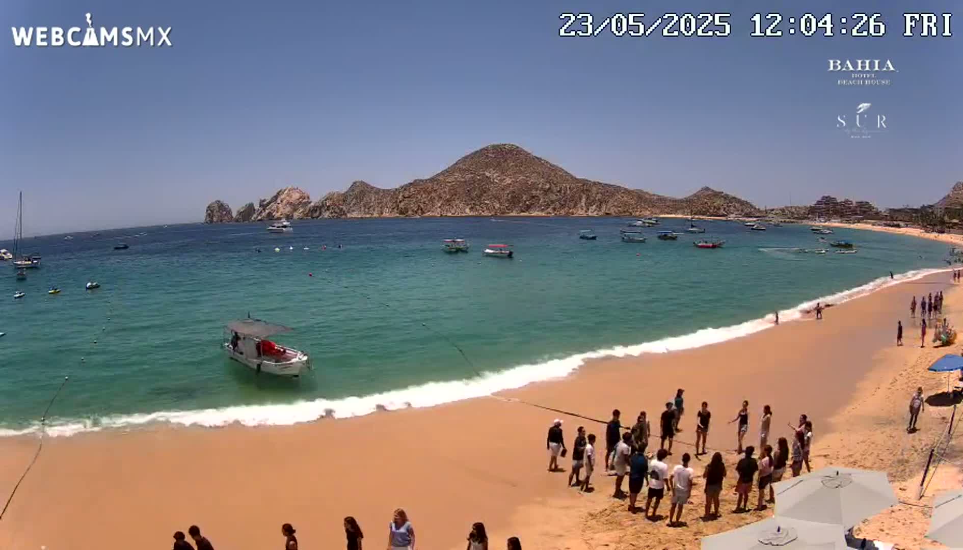 A sandy beach with calm, turquoise water, boats in the bay, and several people gathered on the shore under a sunny sky.