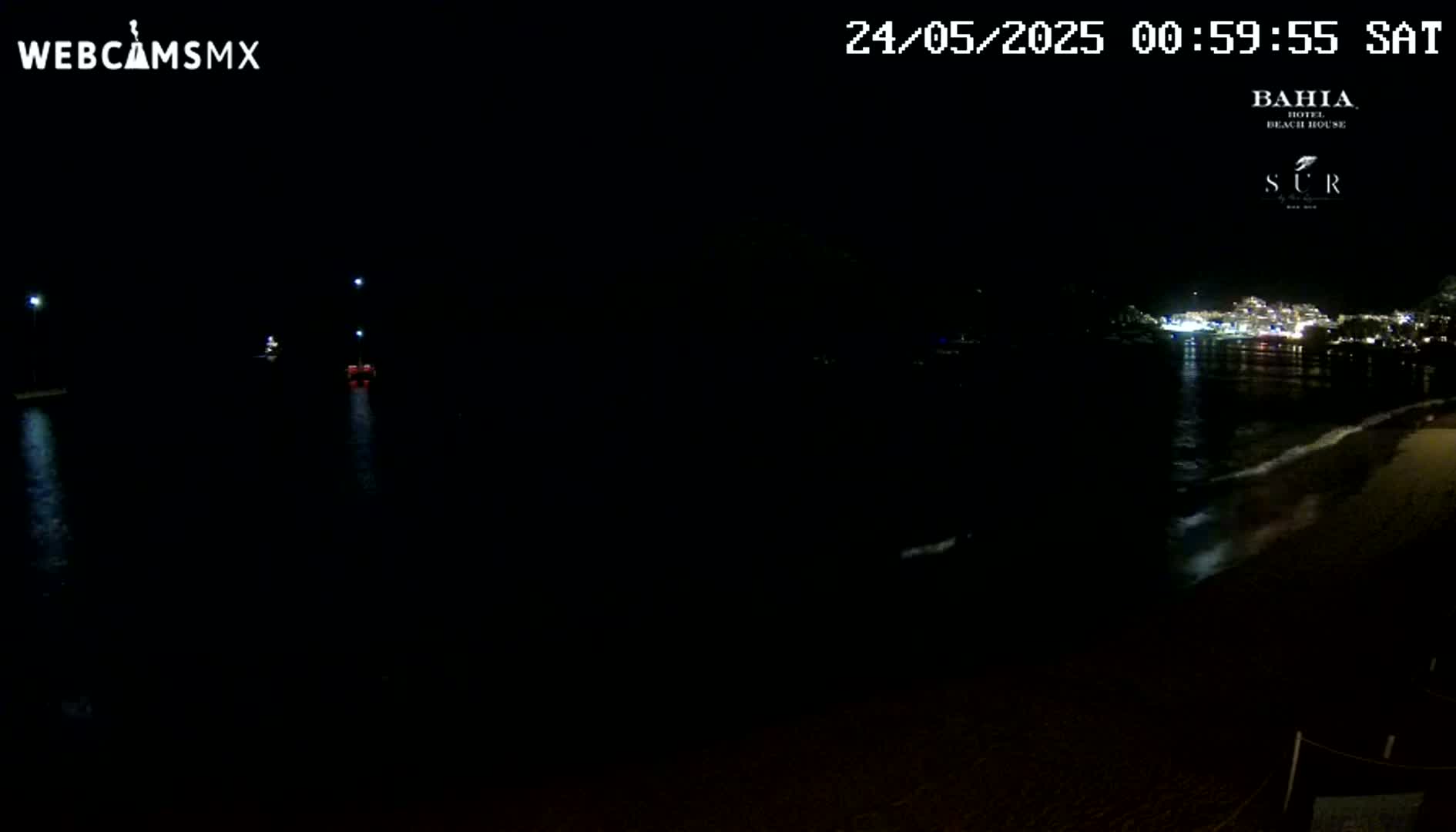 A nighttime view of a dark body of water with a brightly lit town visible in the distance, gentle waves lapping at a nearby shore.