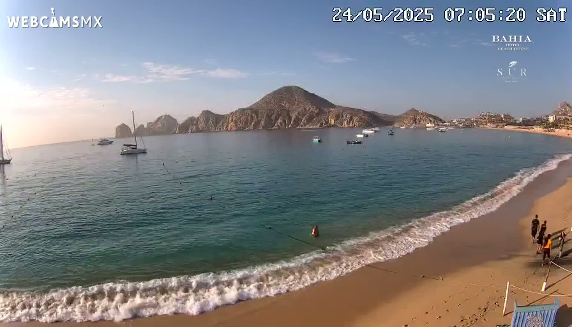 A sandy beach with gentle waves laps the shore, offering a view of a calm bay with several boats and a mountainous peninsula under a clear, sunny sky.