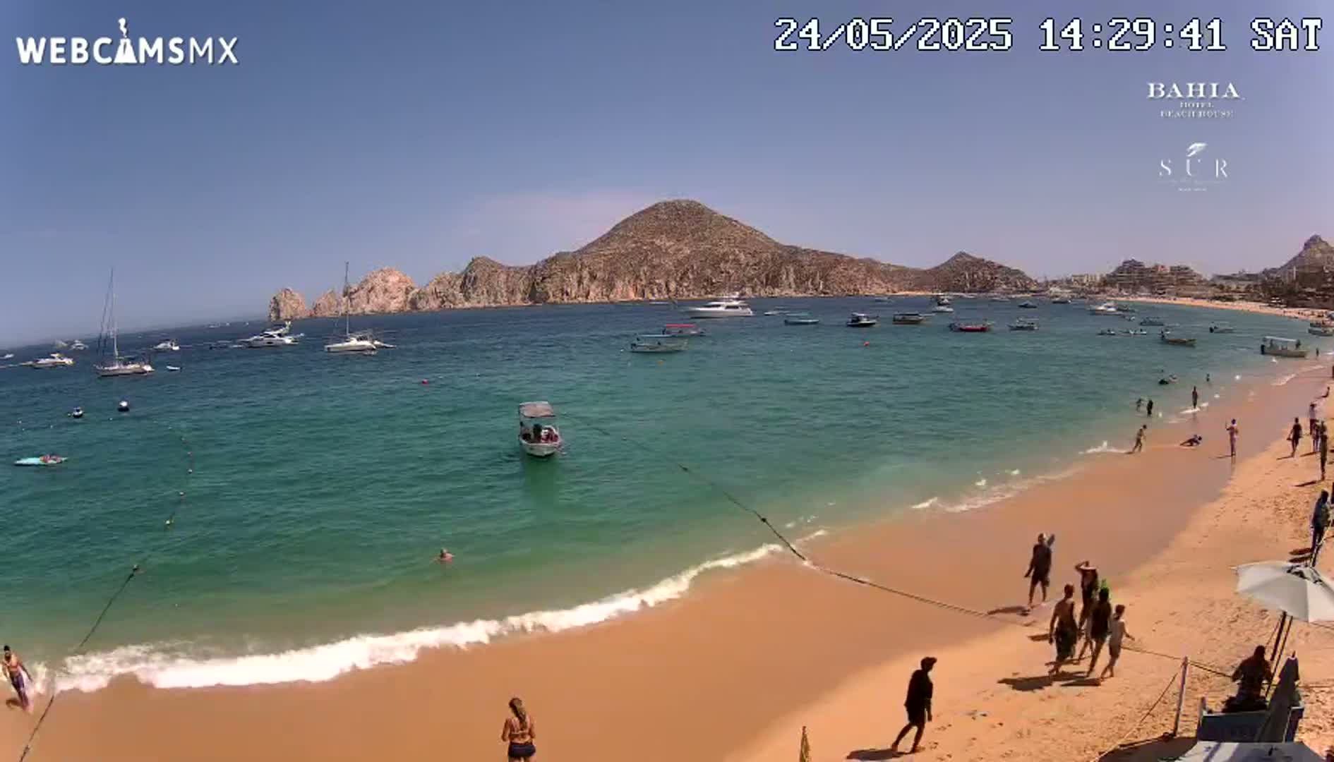 A sandy beach with calm, turquoise water and several boats offshore, under a clear sunny sky, has people swimming in the water and relaxing on the shore.