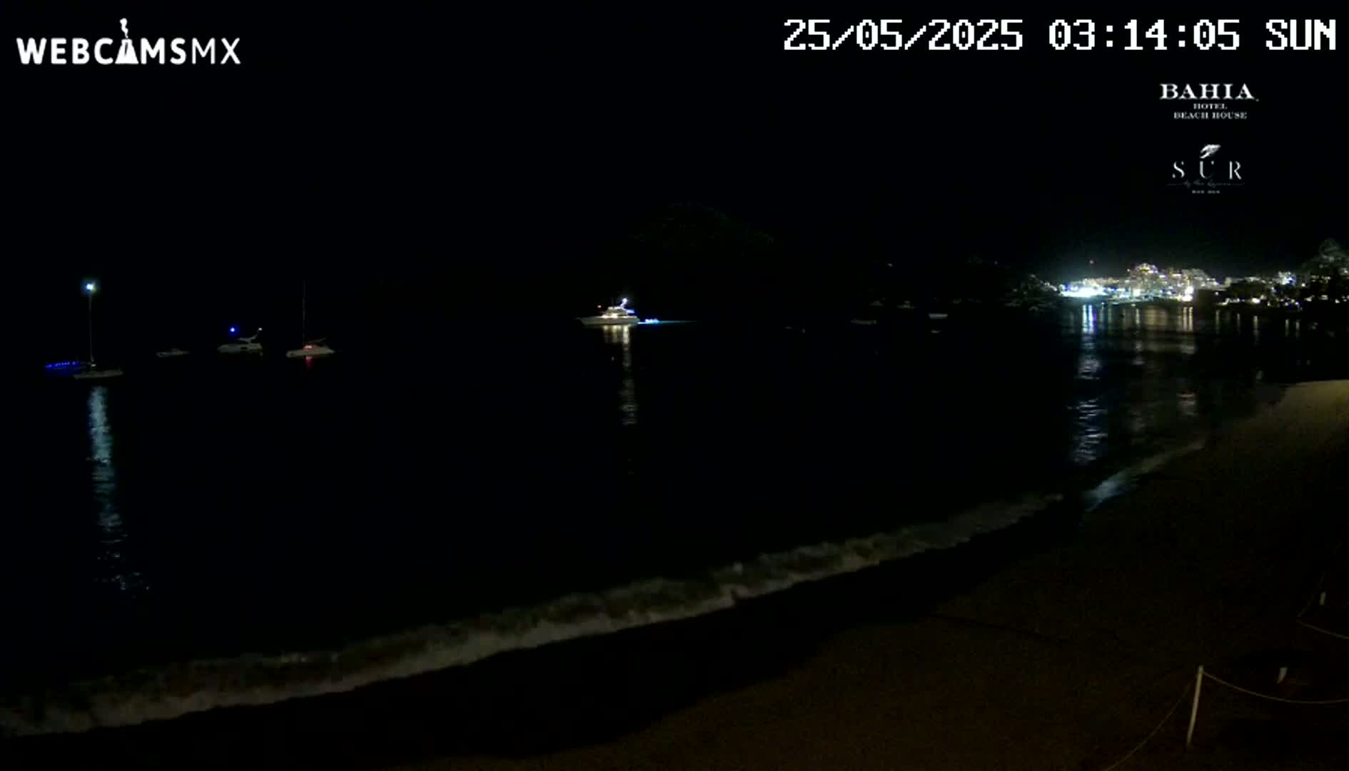 At night, a dark beach with gently lapping waves is shown, with several boats visible on the calm water and lights from buildings across the bay.