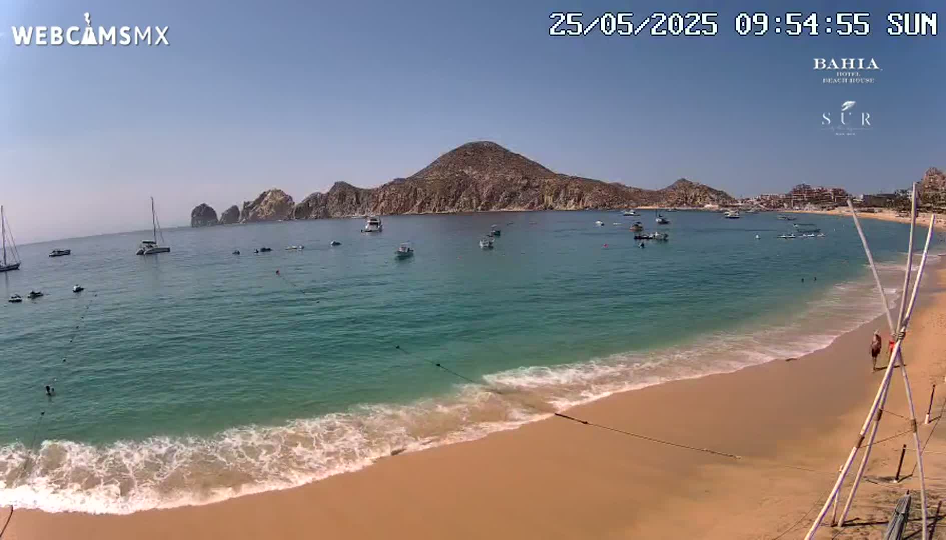A sandy beach with turquoise water laps gently at the shore under a clear blue sky, with several boats visible in the calm bay and a large landmass in the background.