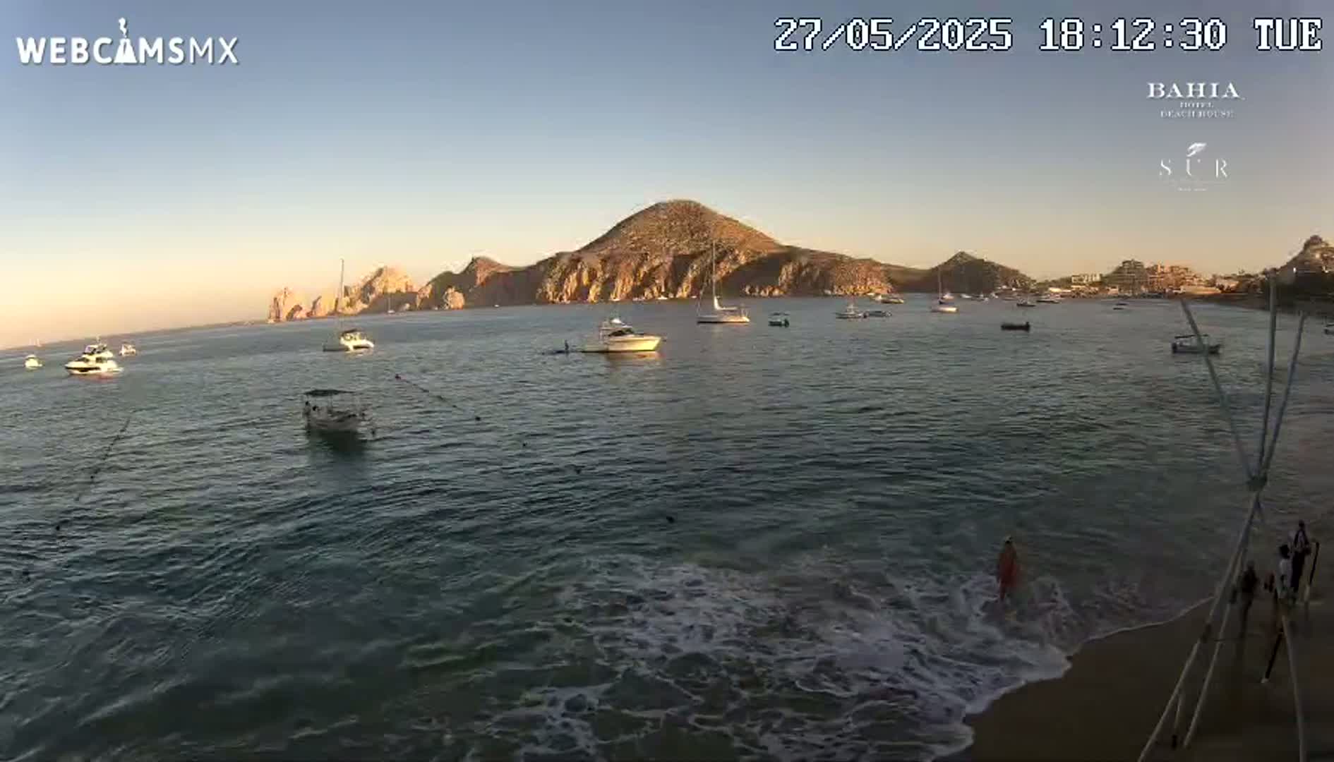 Several boats are anchored in a calm bay at sunset, with a sandy beach and a large landmass in the background.