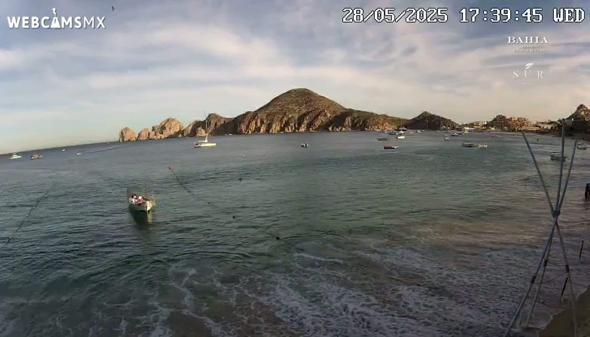 A calm bay with several boats and a large landmass in the background under a partly cloudy sky.