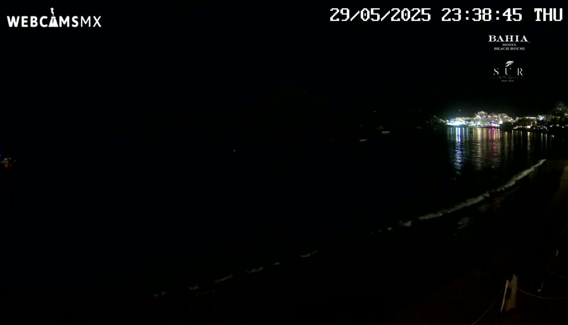 A nighttime beach scene shows a dark body of water reflecting the lights of buildings across the bay, with gentle waves lapping the shore.