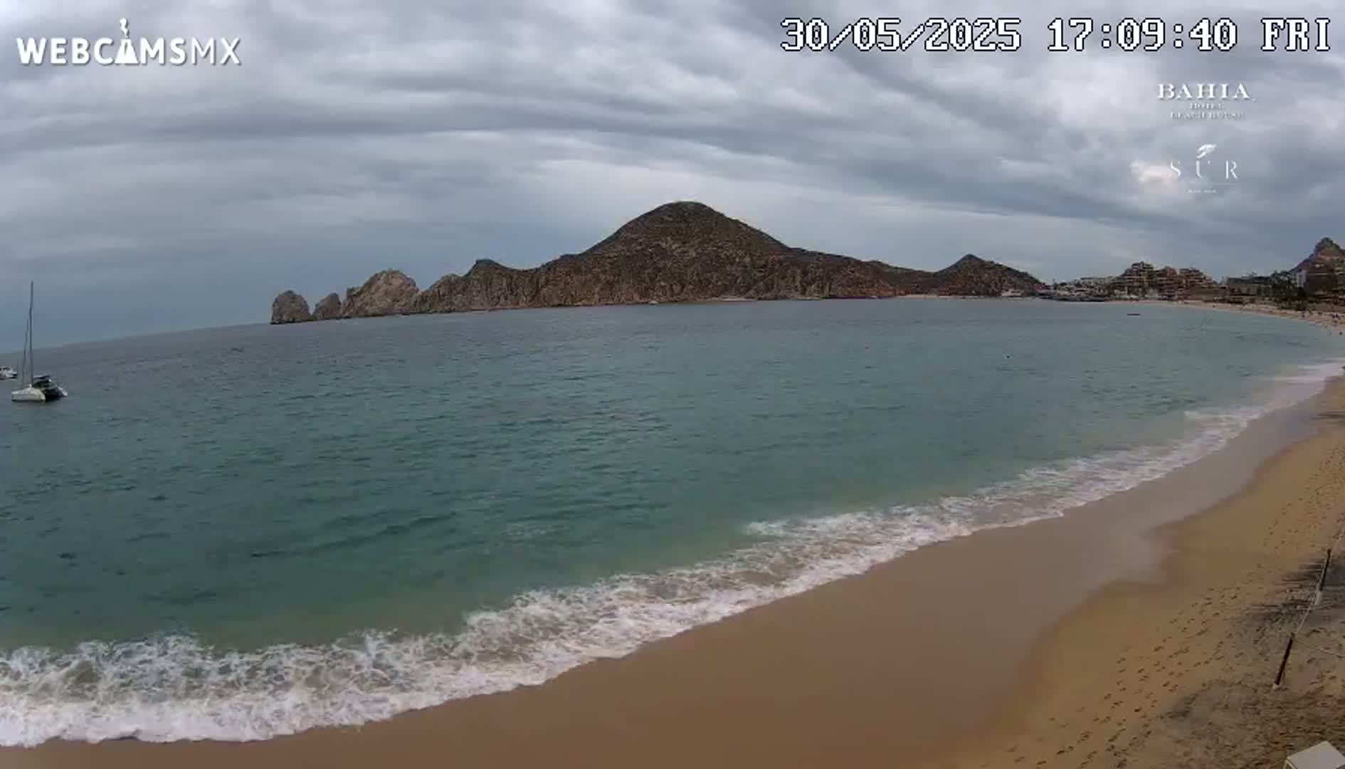 A sandy beach with gentle waves laps against the shore, under an overcast sky, with a large landmass in the distance.