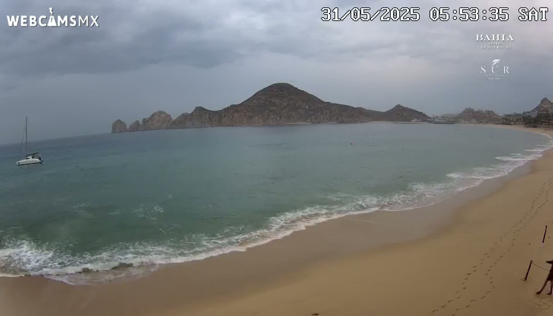 A sailboat is anchored in a calm bay with a sandy beach and a large hill in the background under an overcast sky.