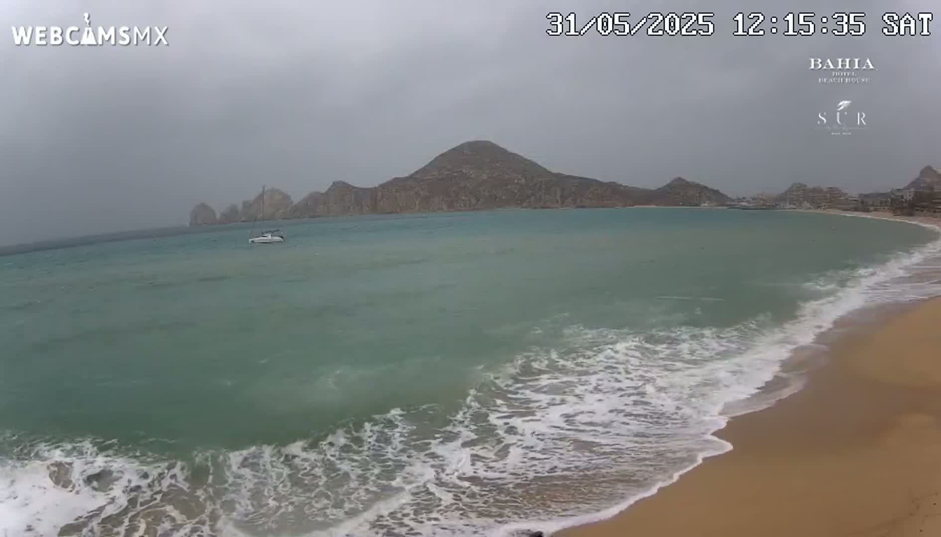A sandy beach with moderate waves laps against the shore under an overcast sky, with a boat visible in the calm water and mountains in the distance.