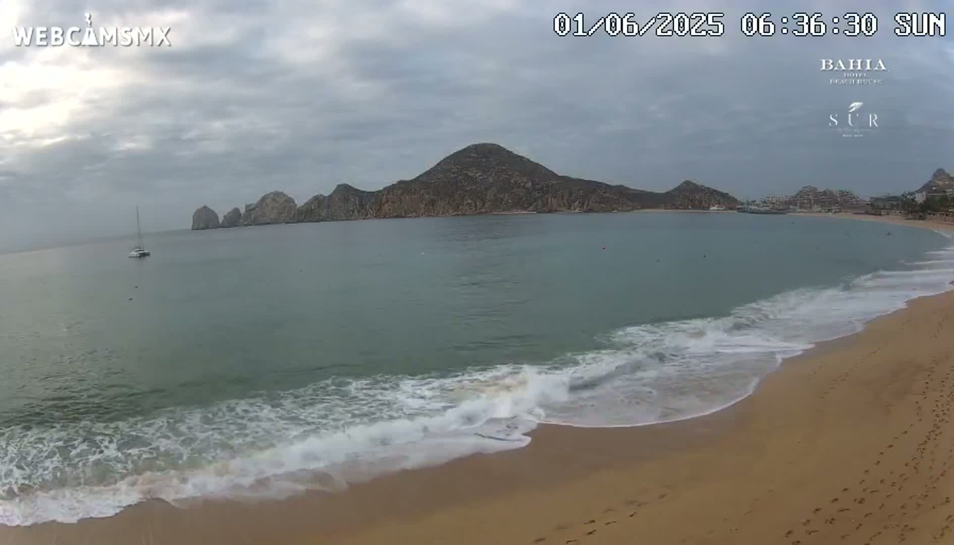 A sandy beach with gentle waves laps the shore under a cloudy sky, with a sailboat visible in the calm bay and a large landmass in the background.