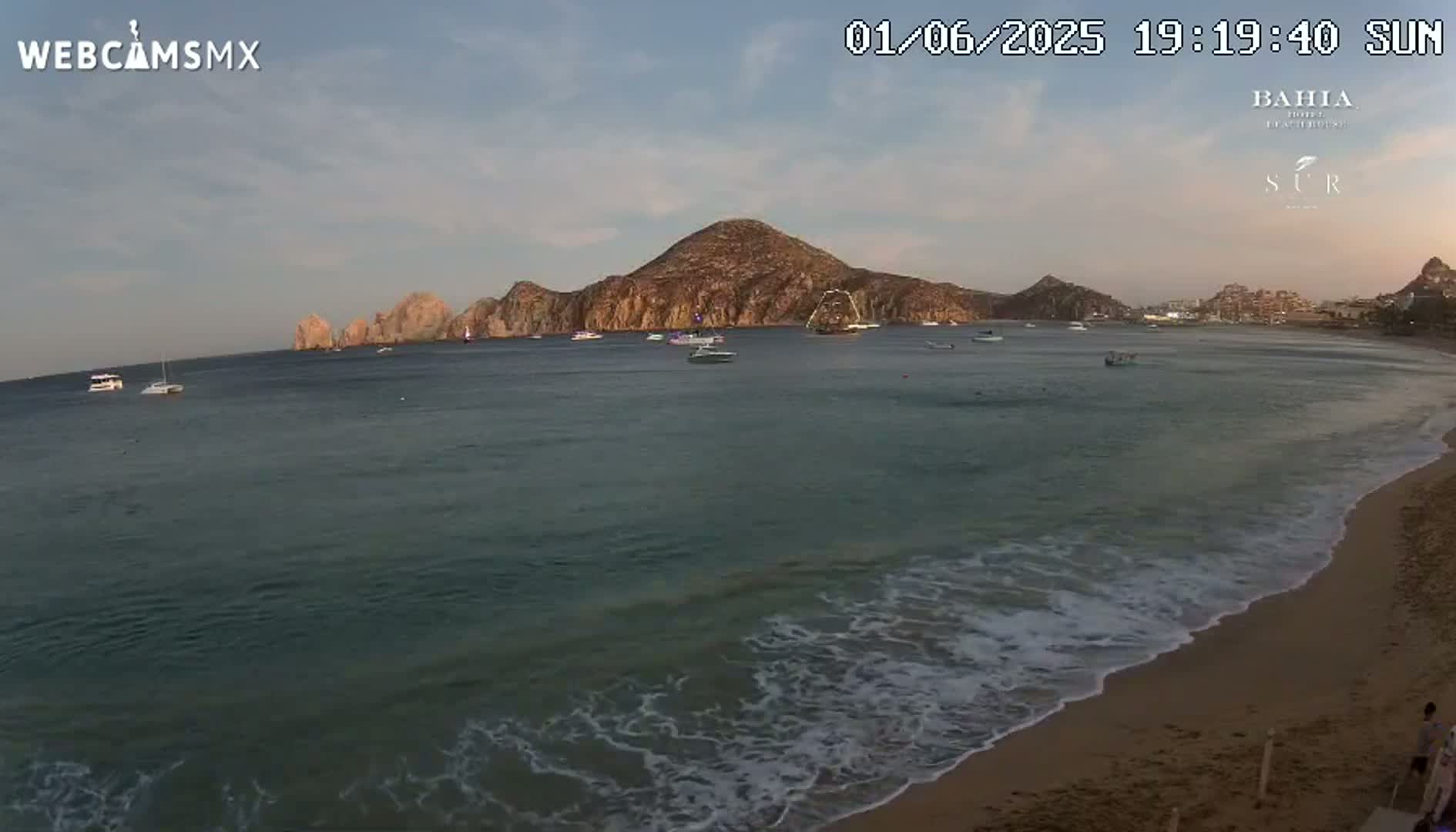 A sandy beach with gentle waves laps the shore under a mostly clear twilight sky, with several boats visible in the calm water and a large landmass in the distance.