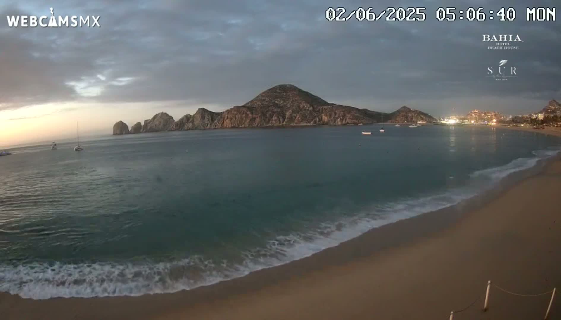 A sandy beach with gentle waves laps the shore under an overcast sky, with a large landmass and several smaller islands visible in the calm water beyond.