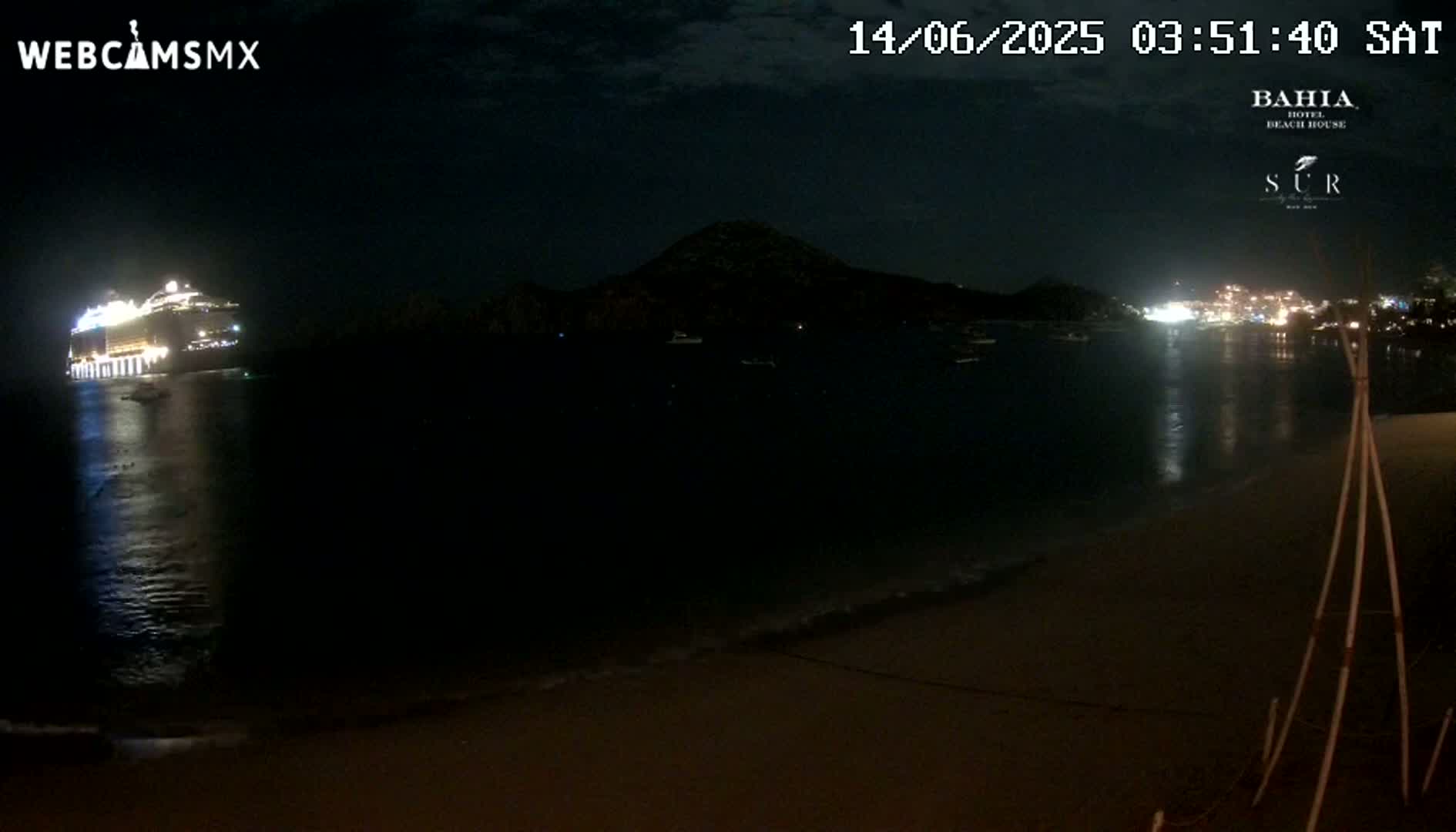A brightly lit cruise ship is in the water at night near a dark shoreline with a hill in the background, under a mostly clear sky.