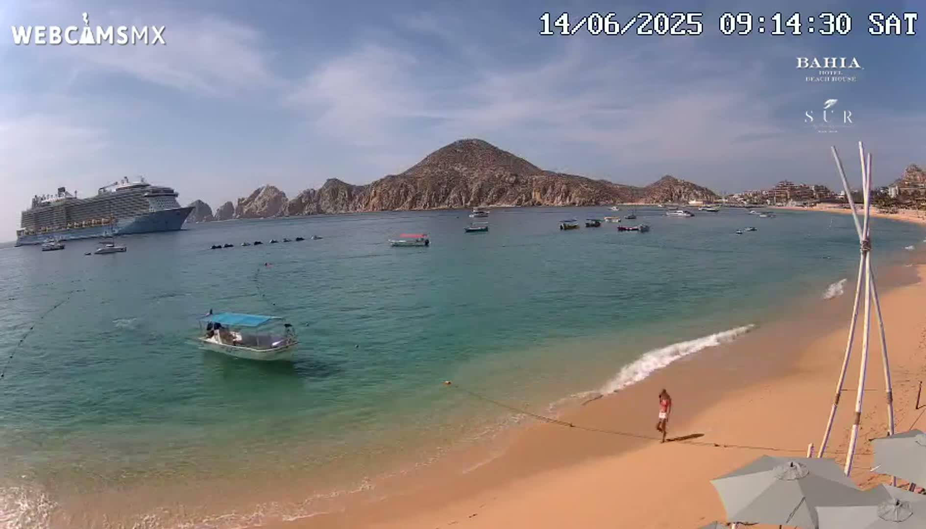 A large cruise ship is anchored in a calm, clear bay near a sandy beach with a person walking along the shore under a mostly sunny sky.