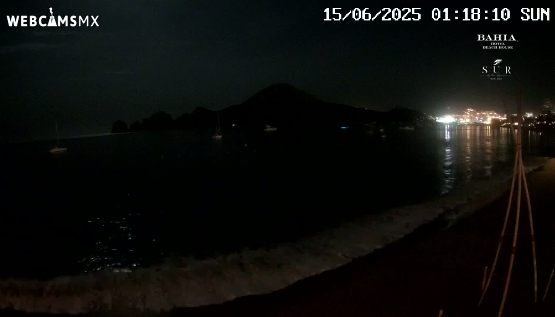 A dark, nighttime beach scene shows gently lapping waves on a dark sandy shore, with a silhouetted mountain and distant lights across the water.