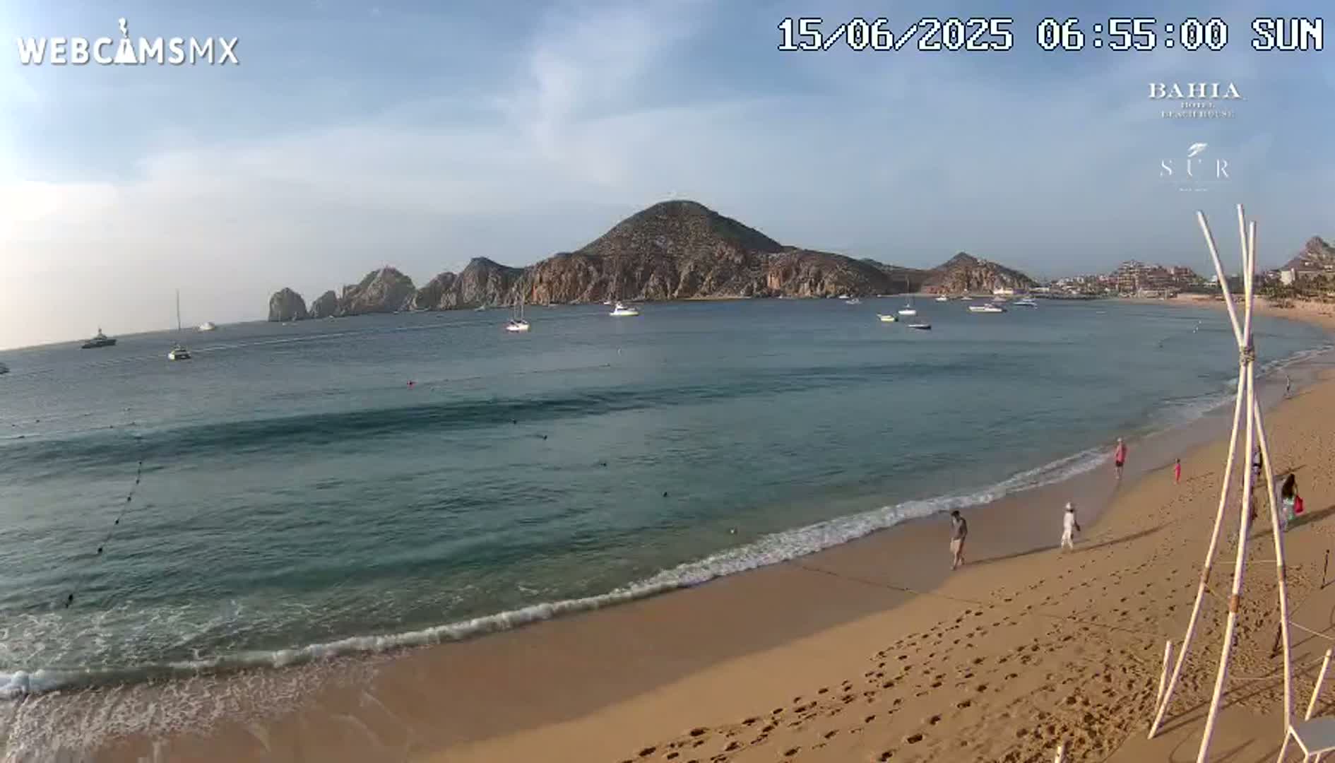 A sandy beach with several people walking along the shore, gentle waves lapping at the sand, and boats visible in the calm, clear water beyond a rocky peninsula under a mostly sunny sky.