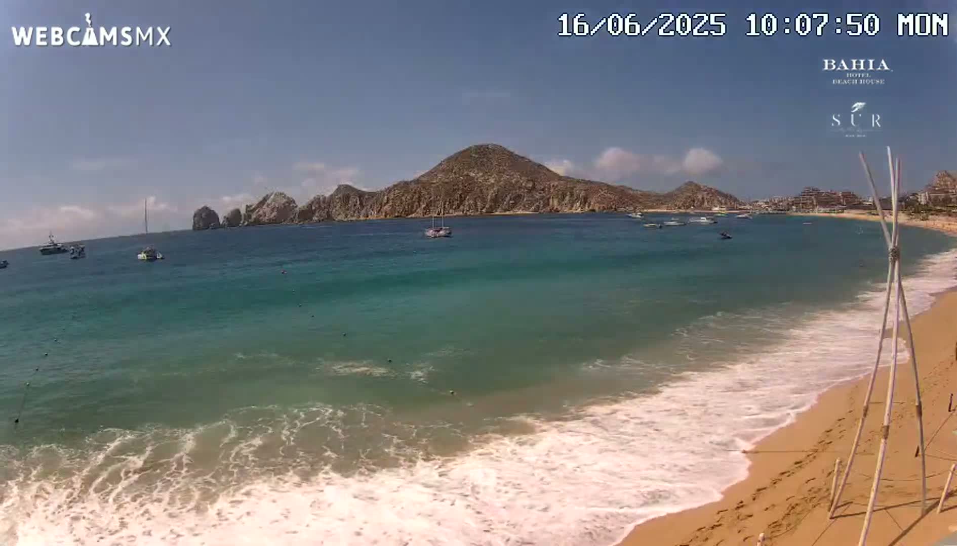 A sandy beach with turquoise water laps gently onto the shore under a clear blue sky, with boats visible on the water and a rocky hill in the distance.