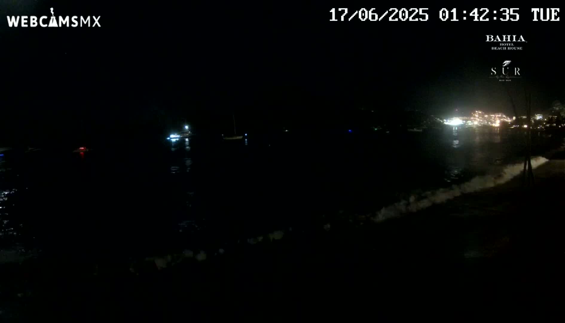 A nighttime view of a dark body of water with small boats and a shoreline with lights in the distance, showing gentle waves lapping the shore.
