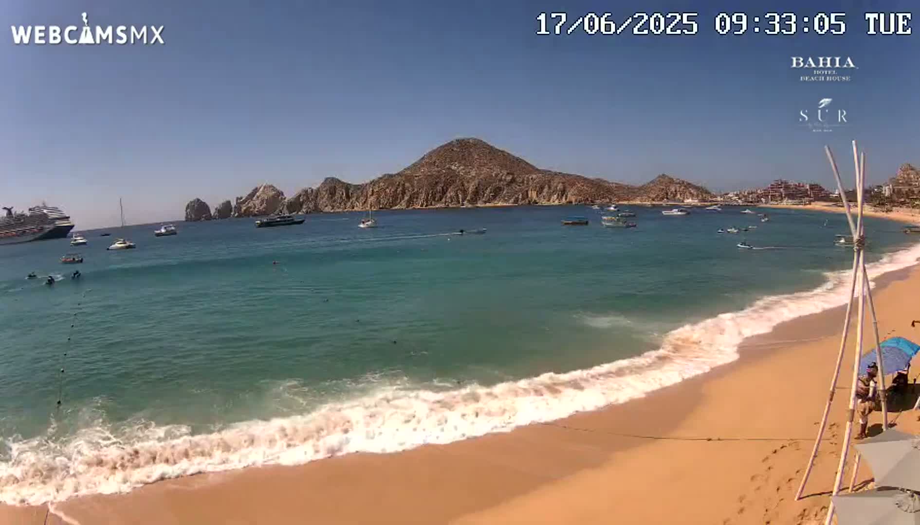 A sandy beach with gentle waves laps the shore under a clear blue sky, with a large body of water containing several boats and a cruise ship in the distance, backed by a mountainous landscape.