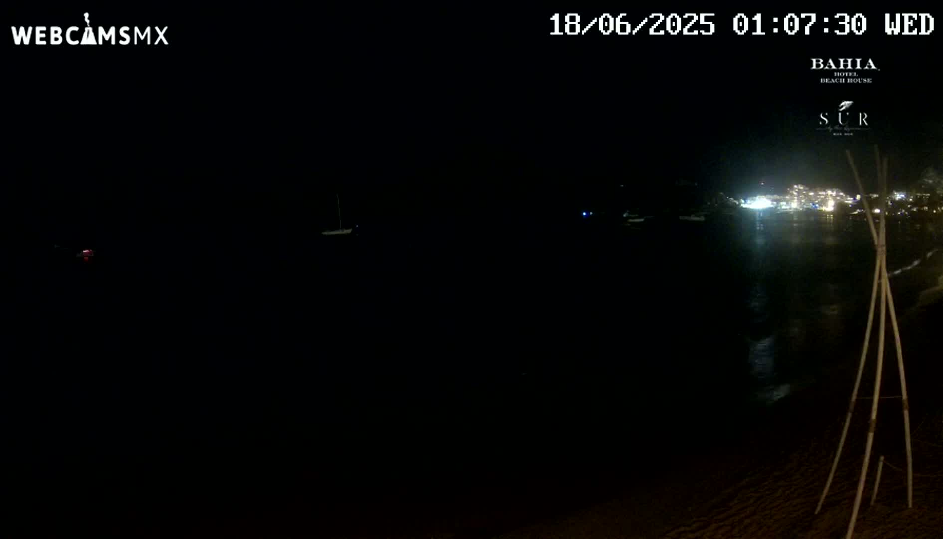 A dark nighttime scene shows a body of water with lights faintly visible in the distance, a dark beach in the foreground, and a tripod-like object on the sand.