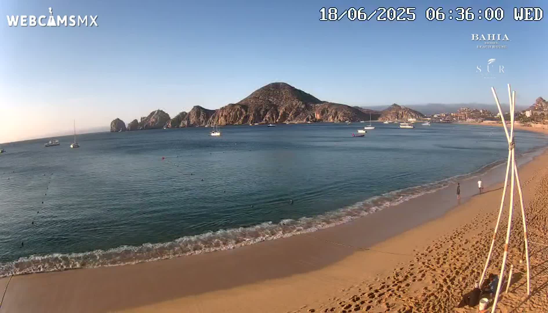 A sandy beach with calm, blue water laps gently at the shore under a clear, bright sky, with boats in the bay and rocky hills in the distance.