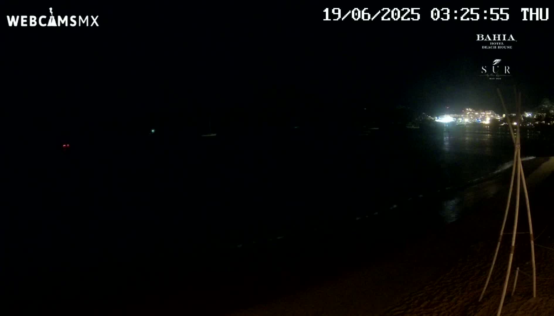 A dark nighttime beach scene shows a calm ocean reflecting the lights of a distant town under a clear sky.